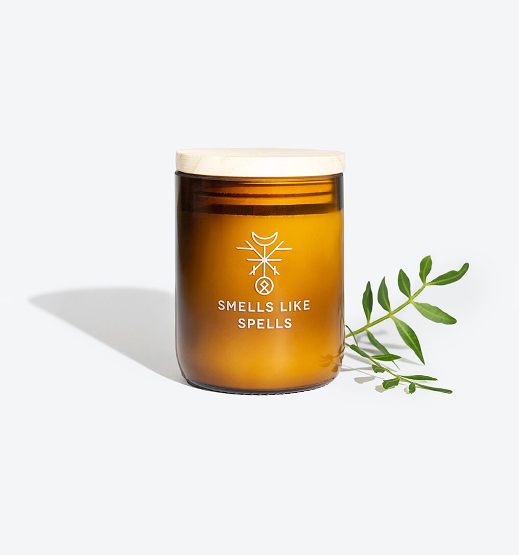 3. Smells Like Spells FRIGGA candle in amber glass jar with wooden lid, styled with green foliage for a natural ambiance
