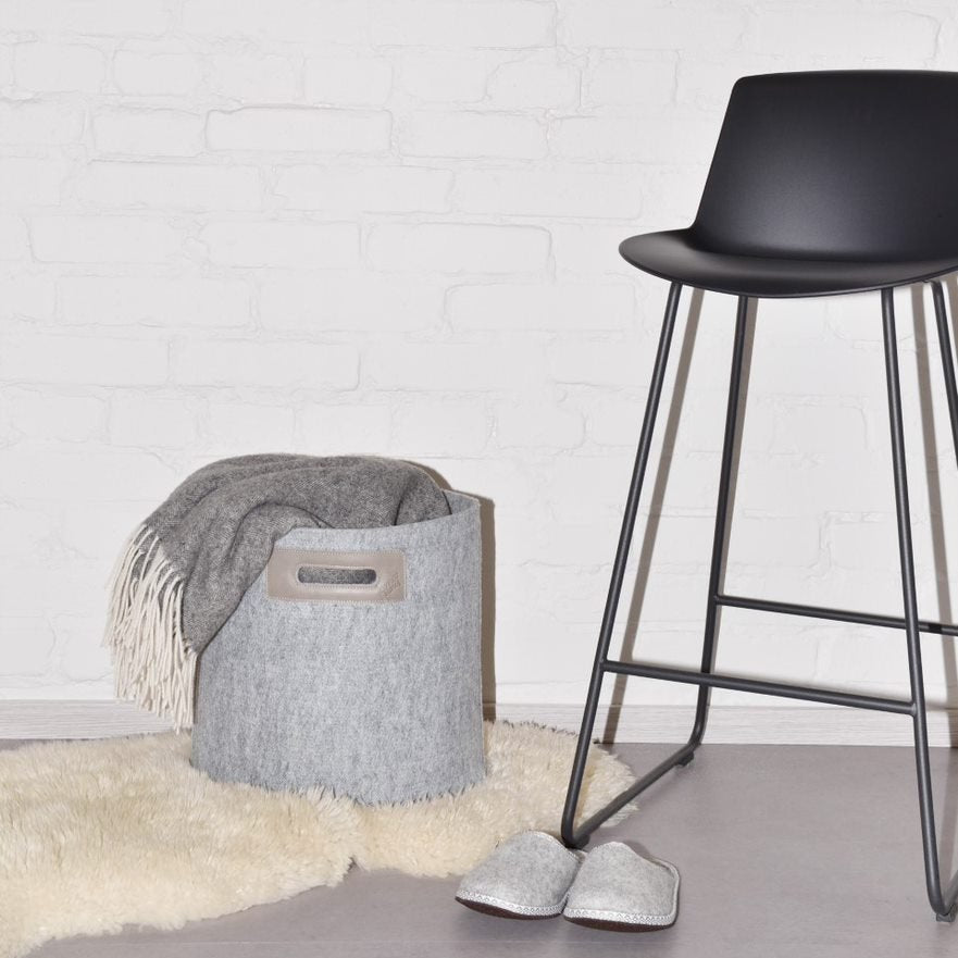 1. Lifestyle image of grey Omaking felt basket with leather handle, styled with a grey blanket and slippers, next to a black chair on a white rug