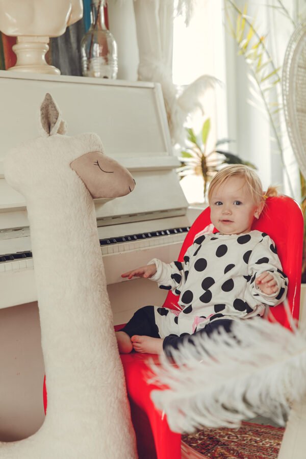 5. Child in polka dot outfit sitting next to beige giant plush alpaca toy by piano