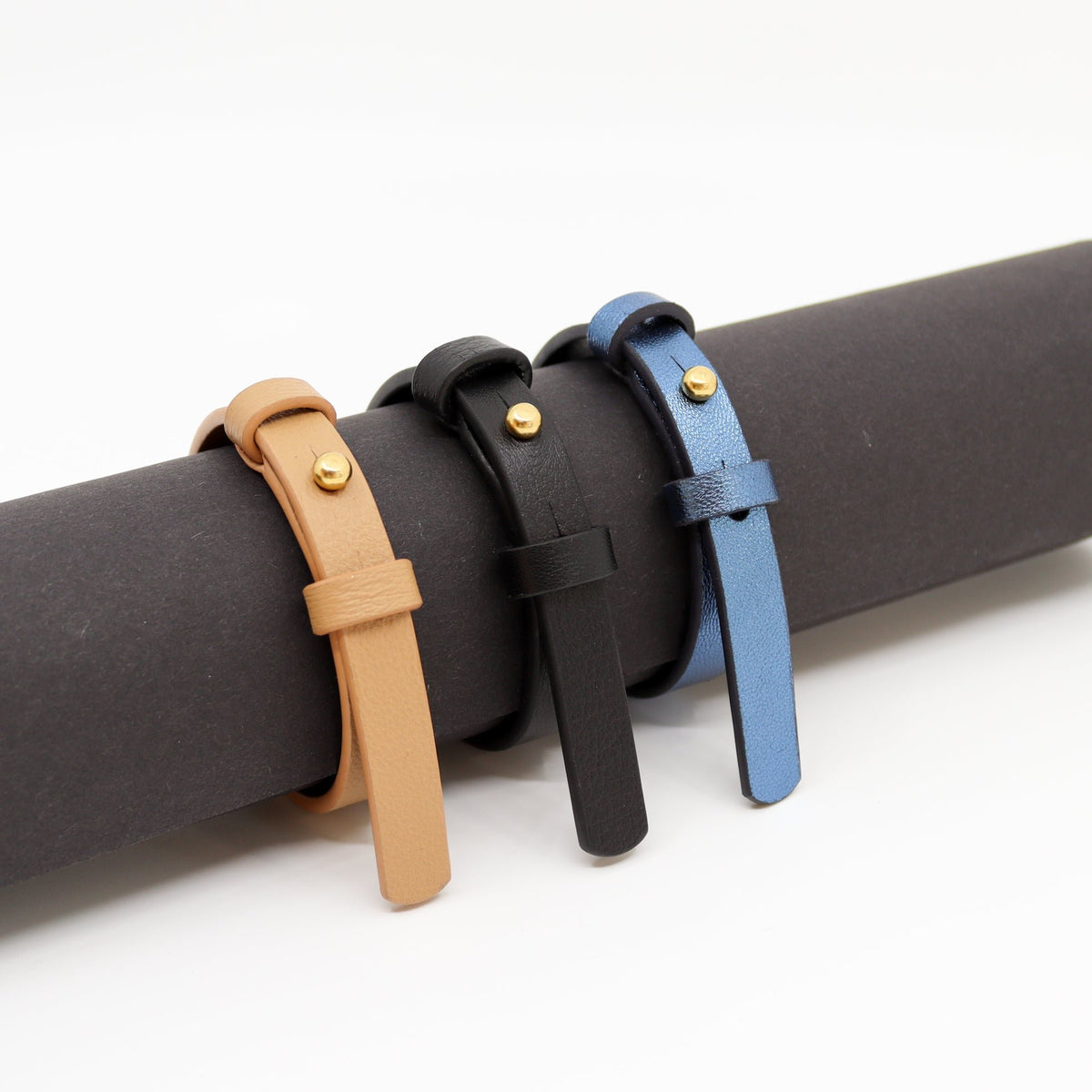 3. Trio of Tairi Roosve leather bracelets in desert tan, black, and blue with brass fasteners, displayed on a roll