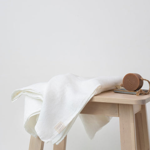4. White waffle linen towel with Linen Tales label draped over wooden bench, accompanied by a natural soap bar, highlighting luxury bath essentials