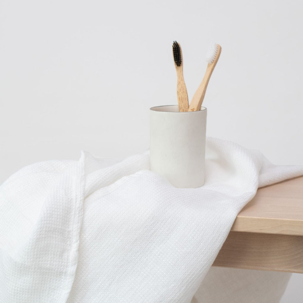 3. White waffle linen towel draped over wooden bench with ceramic cup holding bamboo toothbrushes, emphasizing natural and eco-friendly bathroom decor