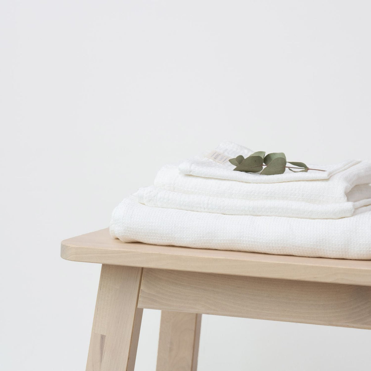 1. Set of white waffle linen towels stacked on a light wooden bench with eucalyptus leaves, showcasing soft texture and minimalist design