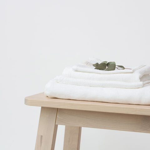 1. Set of white waffle linen towels stacked on a light wooden bench with eucalyptus leaves, showcasing soft texture and minimalist design