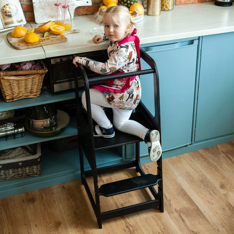 5. Child in floral dress on black Montessori helper tower step stool in kitchen
