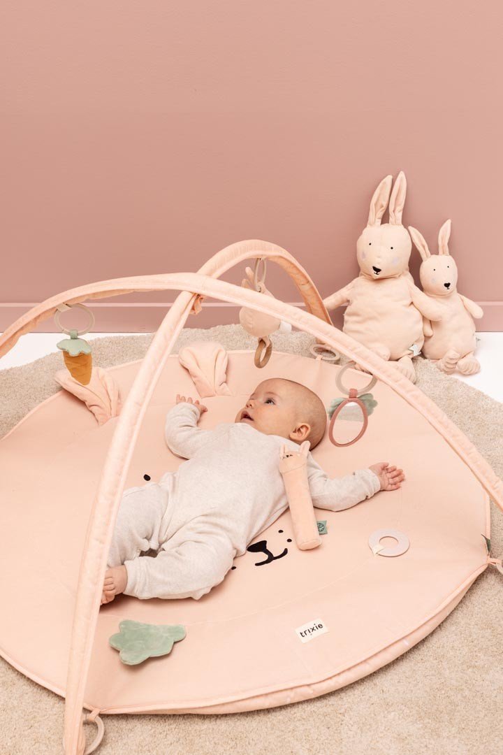 1. Baby lying on Trixie Baby Mrs. Rabbit activity play mat with arches and toys in a pink room