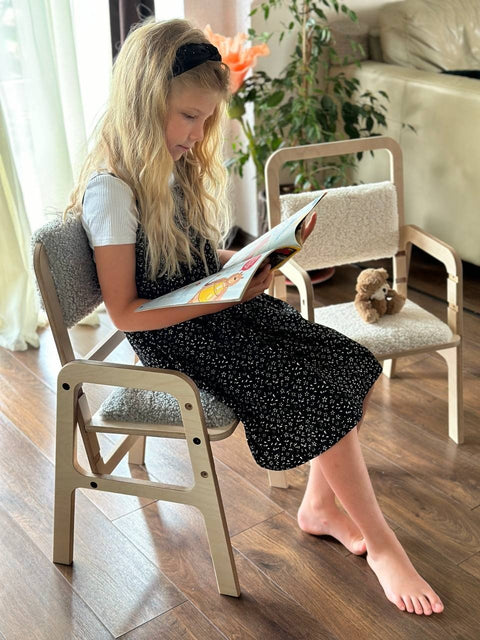1. Young girl reading a book while sitting on an adjustable Luula children's chair with grey padding, in a bright living room