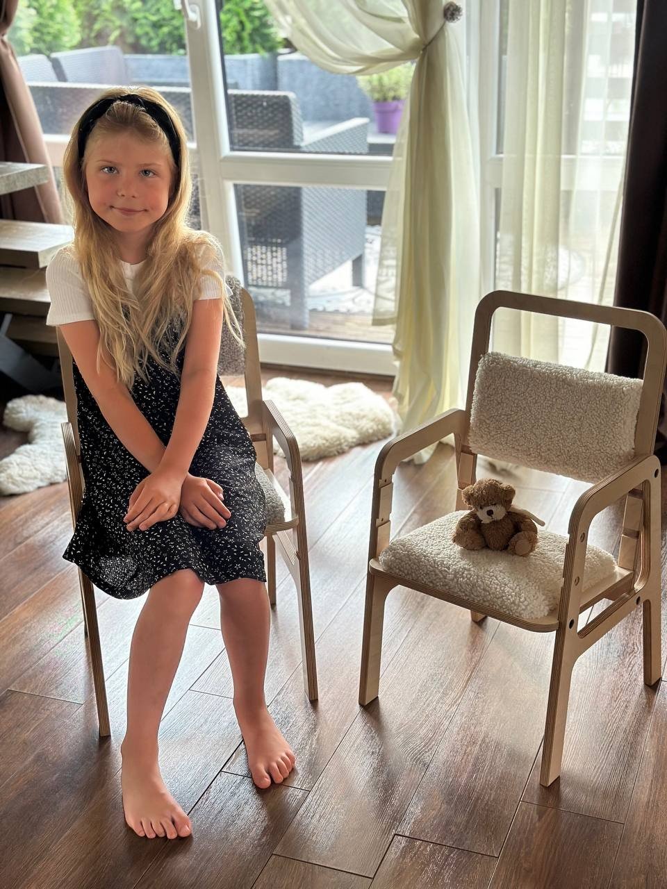 4. Girl sitting next to Luula adjustable chair in natural wood with vanilla lamb padding, in a bright room with large windows
