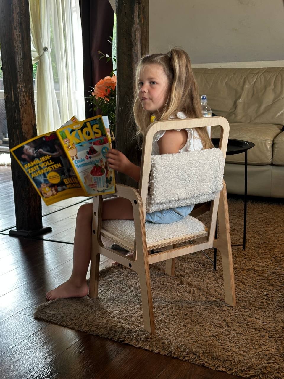 3. Girl sitting on Luula adjustable chair in natural wood with vanilla lamb padding, reading a magazine in a cozy room