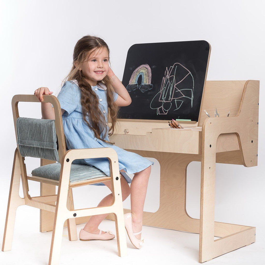 1. Girl in blue dress sitting at Luula mint green Montessori table and chair set, drawing on blackboard in bright room