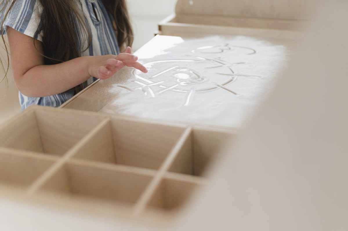 3. Close-up of child's hand drawing on sand surface of Luula Montessori table with storage compartments