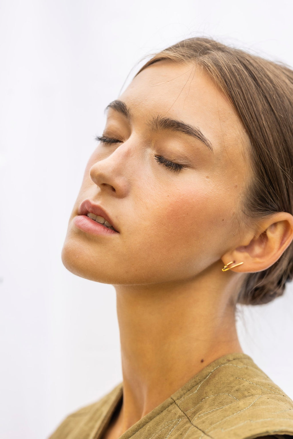 1. Woman wearing Lentsius Sunlit gold-tone earrings with eyes closed, showcasing elegant design against light background