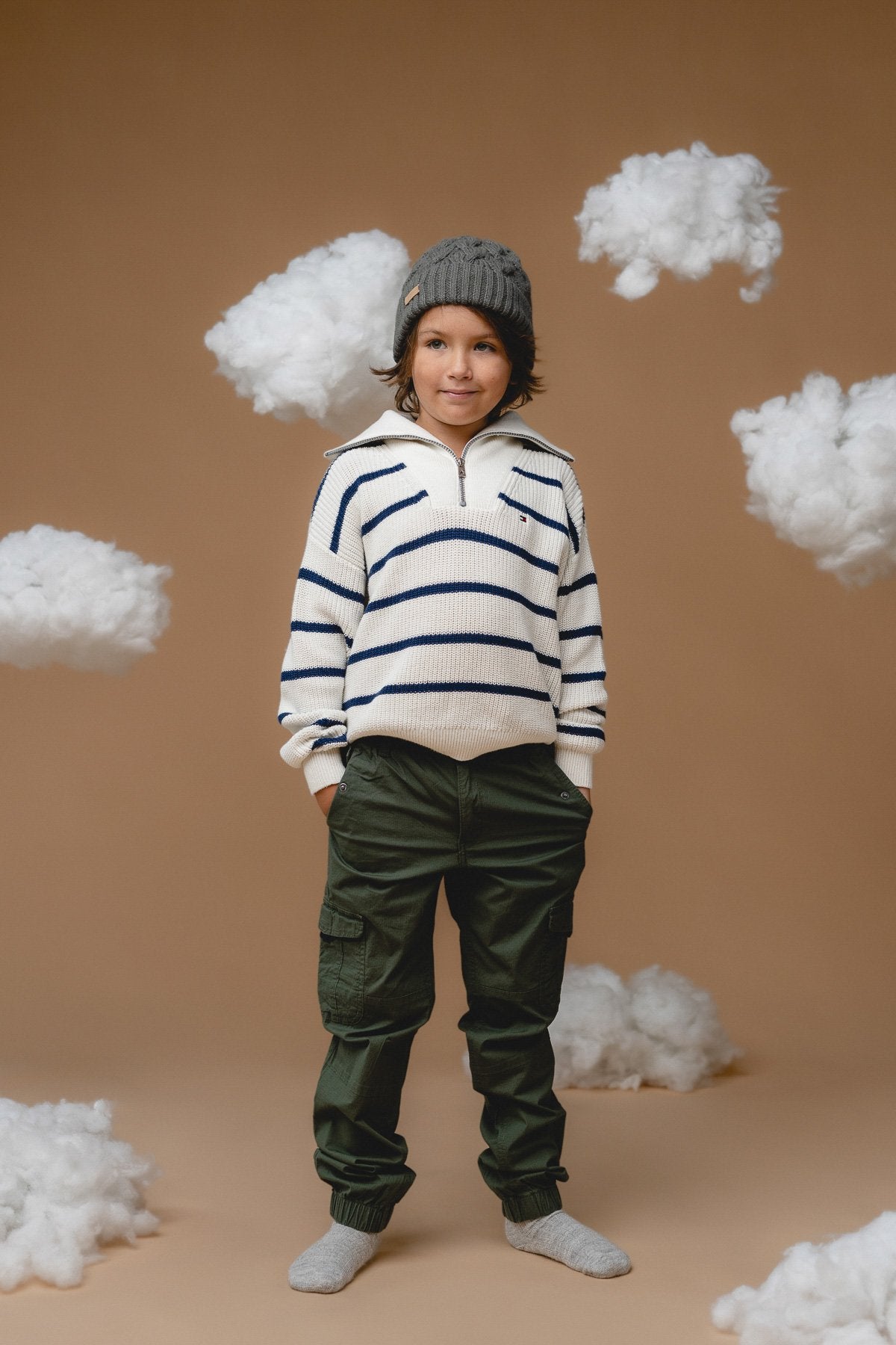 1. Child wearing Alpaka 100% baby alpaca cable hat in botanical green, styled with striped sweater and cargo pants, surrounded by cloud props