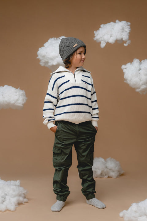 3. Child in Alpaka botanical green baby alpaca cable hat, shown in profile with striped sweater and cargo pants, cloud props setting