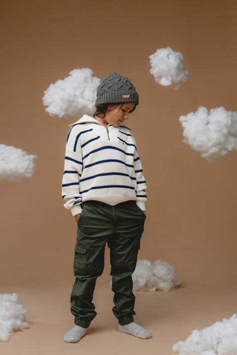 2. Side view of child in Alpaka botanical green baby alpaca cable hat, paired with striped sweater and cargo pants, cloud props in background