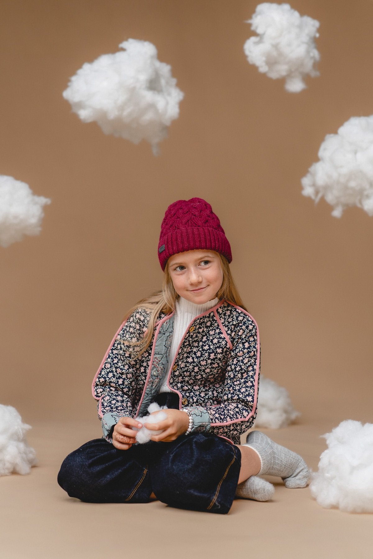 1. Girl wearing Alpaka 100% baby alpaca cable hat in mulberry, surrounded by fluffy clouds, showcasing cozy and stylish design for kids