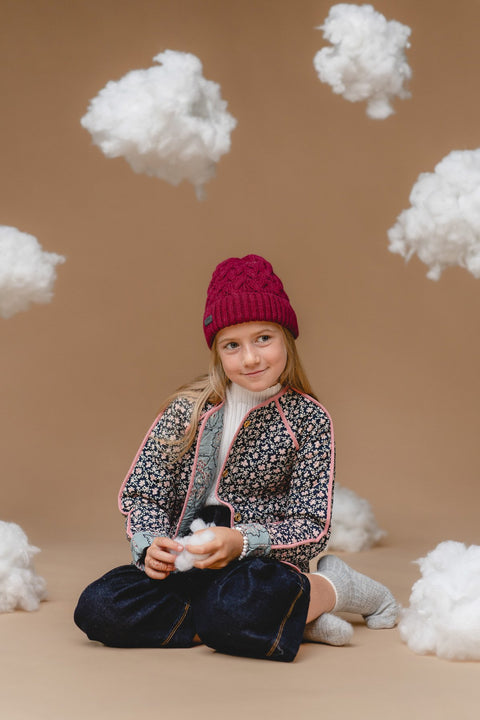 1. Girl wearing Alpaka 100% baby alpaca cable hat in mulberry, surrounded by fluffy clouds, showcasing cozy and stylish design for kids