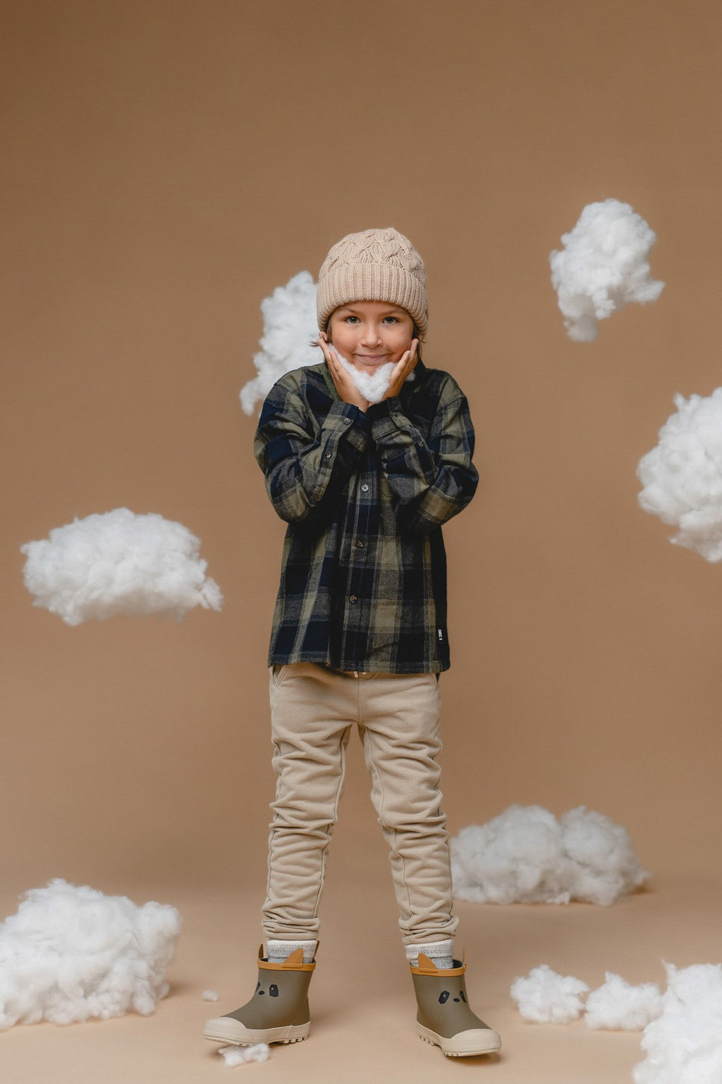 2. Kid in Alpaka wheat-colored baby alpaca cable hat, smiling with hands on cheeks, wearing plaid shirt and beige pants