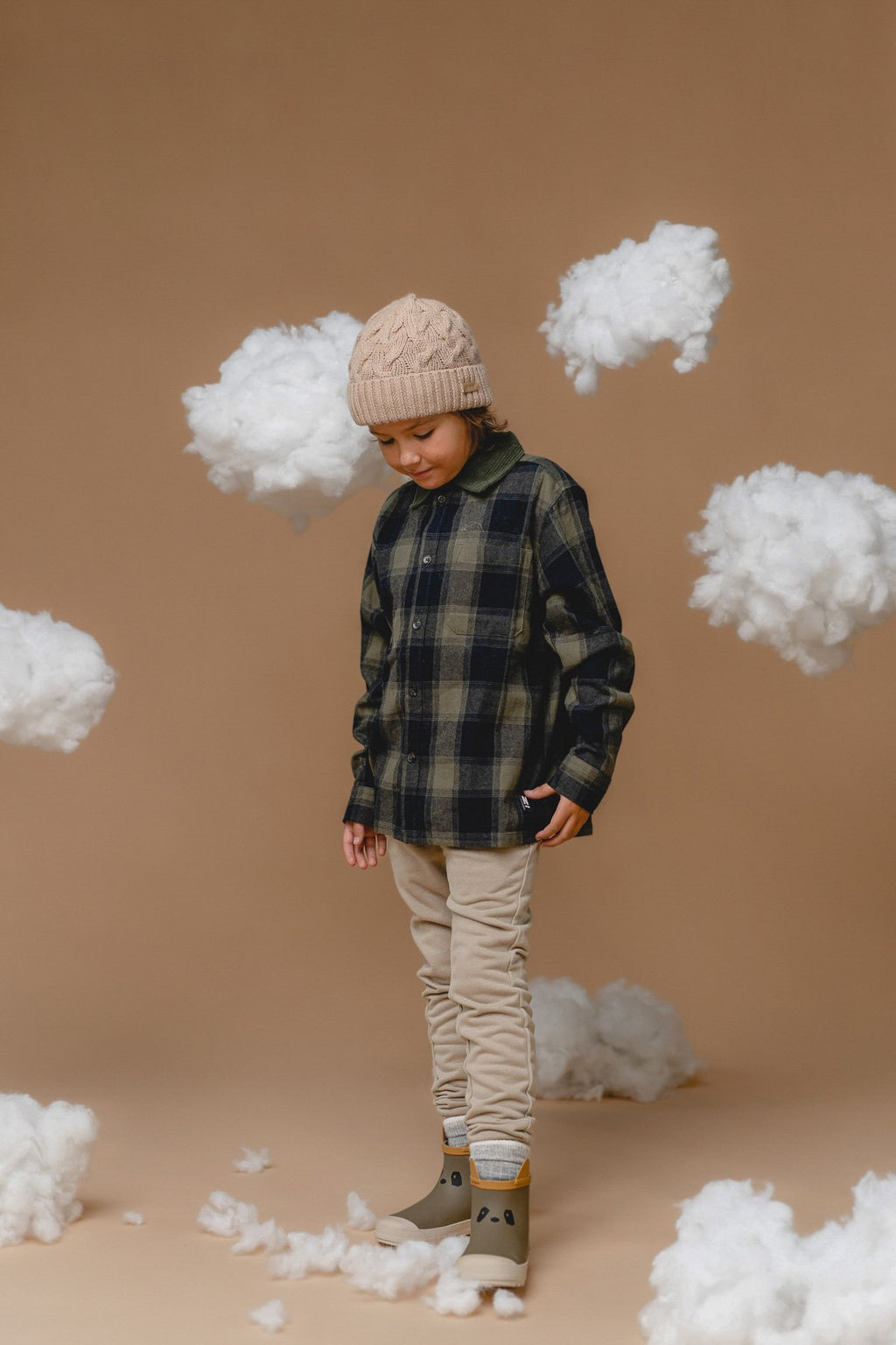 1. Child wearing Alpaka 100% baby alpaca cable hat in wheat, styled with plaid shirt and beige pants, surrounded by fluffy clouds