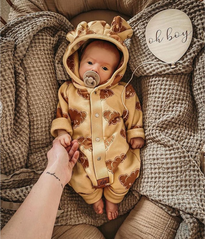 6. Baby lying down wearing yellow jumpsuit with cloud print and hood with ears, holding adult's hand