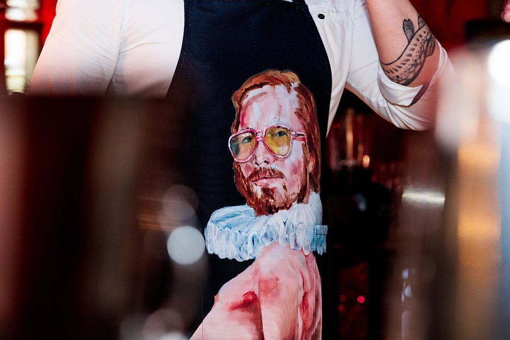 2. Close-up of man wearing Katrin Valgemäe's 'Pervert in the Kitchen' apron with pop art design, in a dimly lit setting