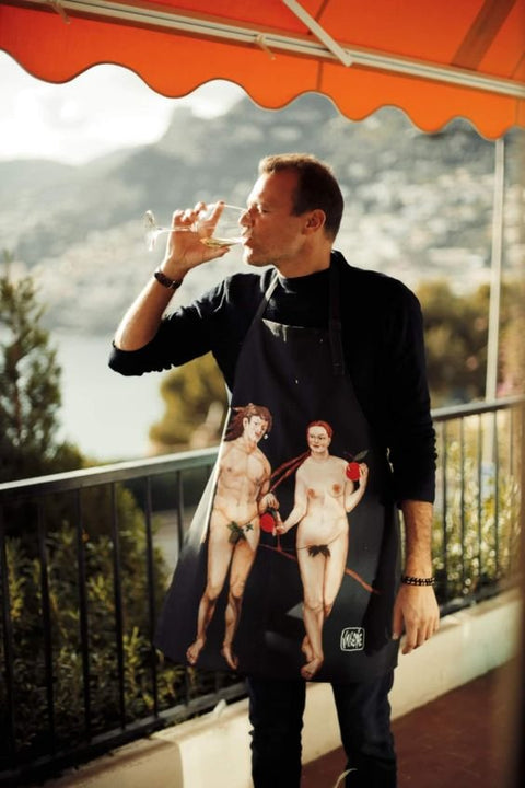 1. Man wearing Katrin Valgemäe apron with Adam and Eve print, standing on balcony with scenic view