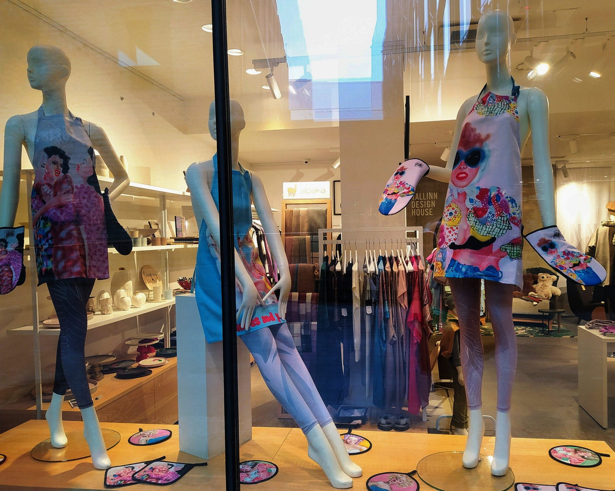 3. Three mannequins in a store window wearing Katrin Valgemäe aprons with artistic prints, surrounded by matching accessories in a contemporary setting
