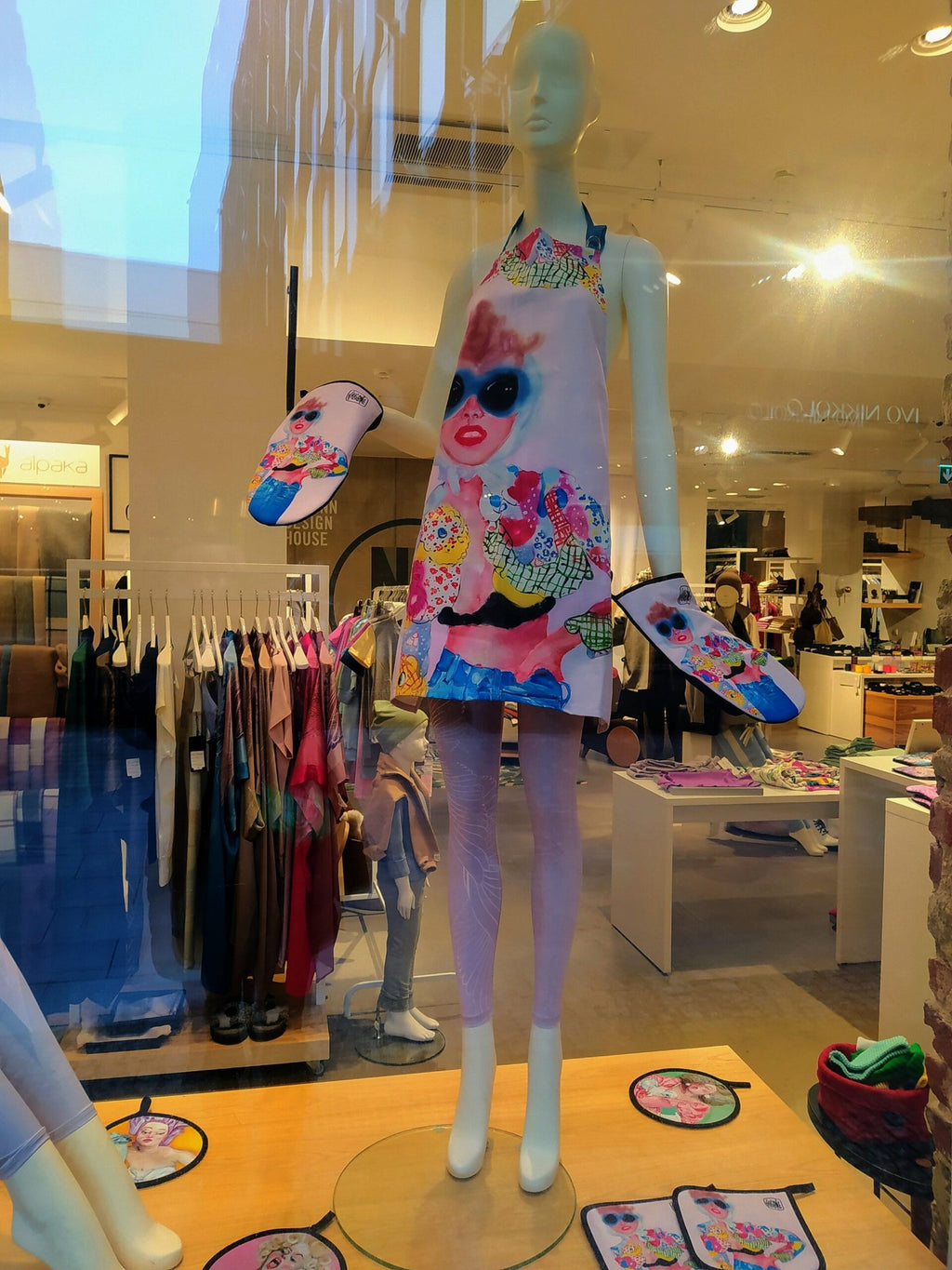 1. Mannequin in store window wearing Katrin Valgemäe apron with colorful pop art design and matching oven mitts