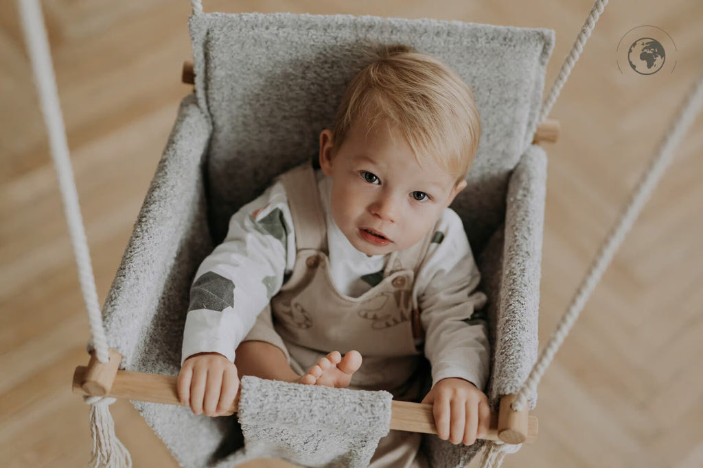 2. Toddler sitting in grey bouclé fabric baby swing with oak wood poles, looking up indoors