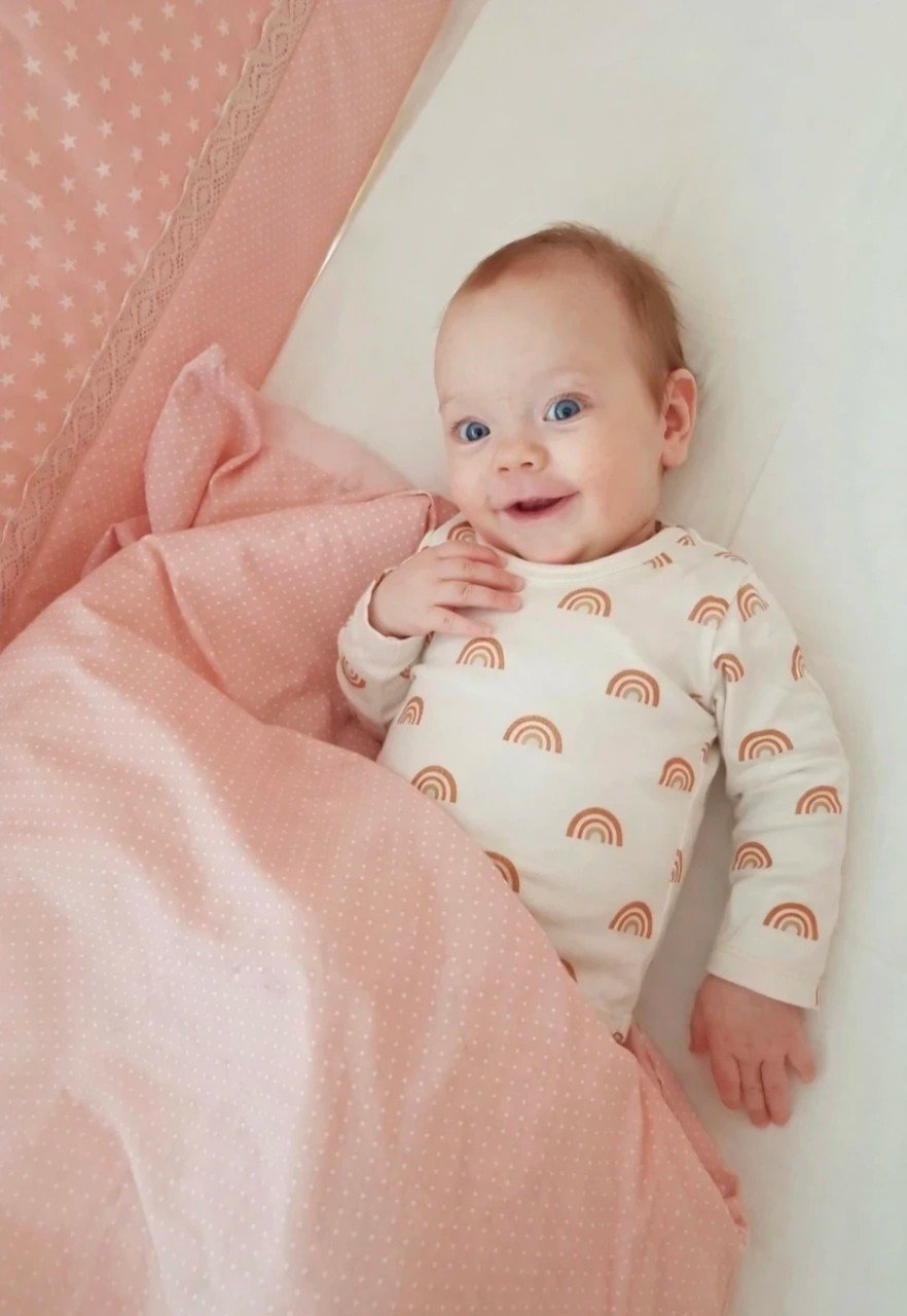 1. Smiling baby wearing Karloova rainbow print bodysuit lying on pink bedding