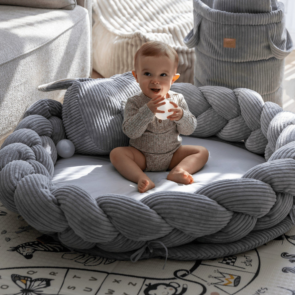 1. Infant sitting in grey braided baby nest play mat by MeowBaby, surrounded by soft corduroy and cotton materials, in a cozy living room setting