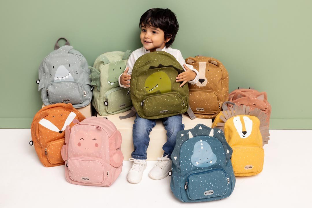 1. Child surrounded by various Trixie Baby backpacks, including Mr. Bear, sitting on a white surface against a green wall