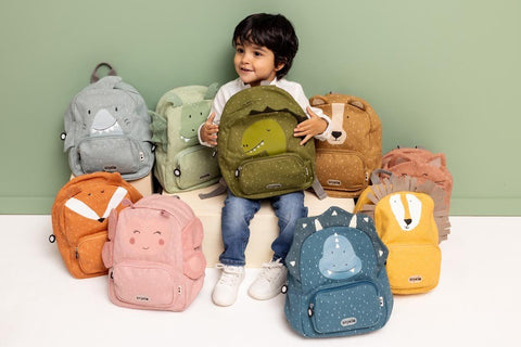 1. Child surrounded by various Trixie Baby backpacks, including Mr. Bear, sitting on a white surface against a green wall