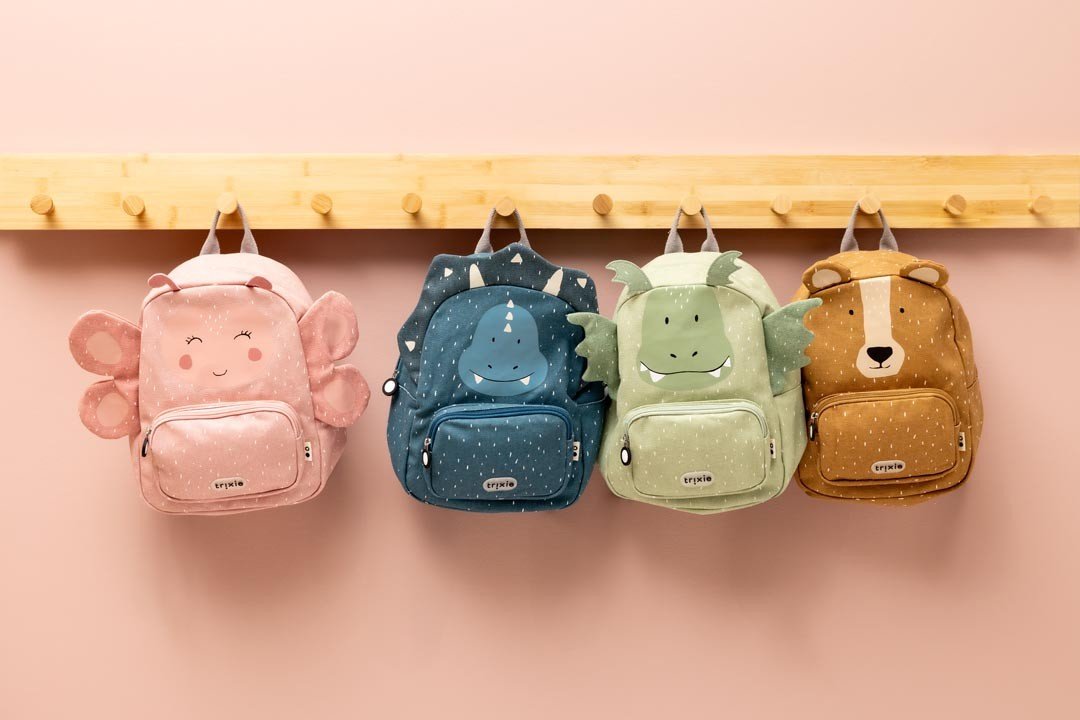 4. Four Trixie Baby backpacks hanging on a wooden rack, including Mr. Bear and Mr. Triceratops, against a pink wall