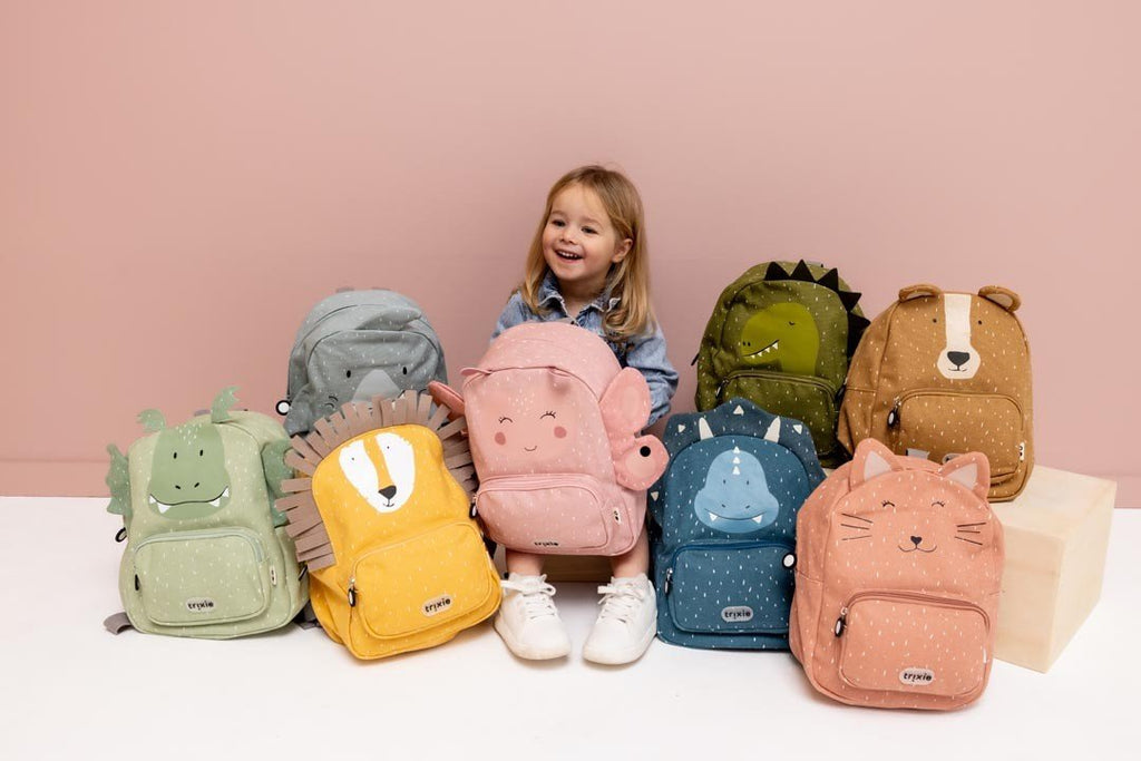 1. Girl smiling with Trixie Baby backpacks, including Mr. Bear, arranged in front of her against a pink background