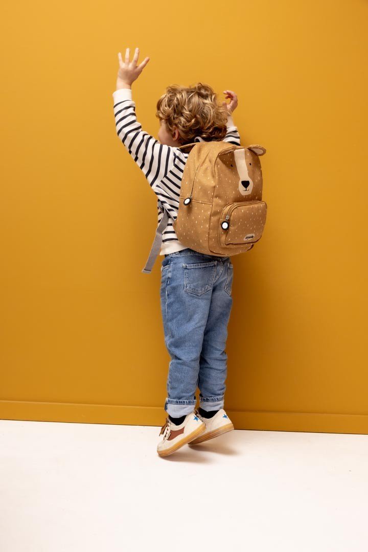 1. Child wearing Trixie Baby Mr. Bear backpack, reaching up against a yellow wall