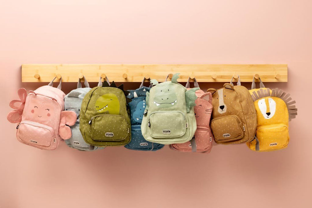 4. Multiple Trixie Baby backpacks, including Mr. Bear, hanging on a wooden rack against a pink wall