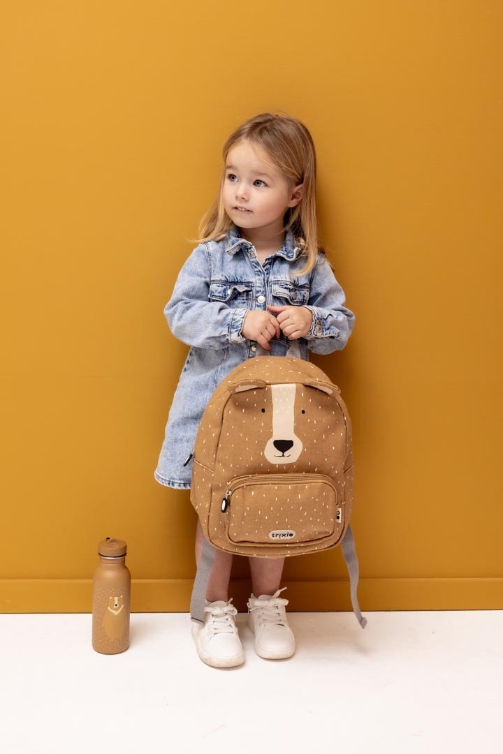 1. Girl holding Trixie Baby Mr. Bear backpack, standing against a yellow wall with matching water bottle