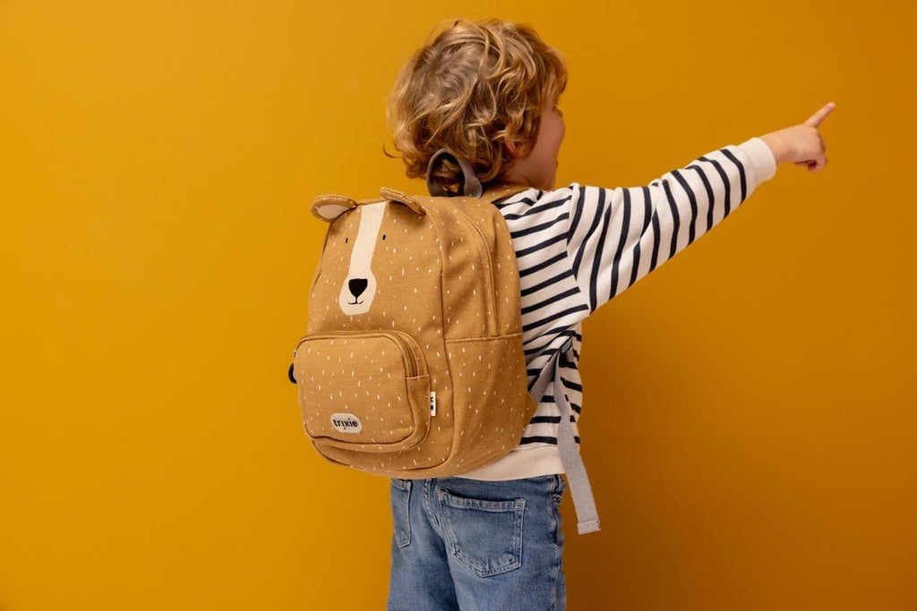 1. Child wearing Trixie Baby Mr. Bear backpack, pointing against a yellow wall