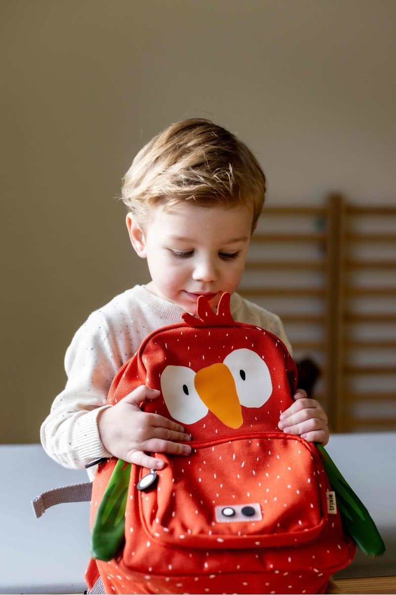 1. Child holding Trixie Baby Mr. Parrot backpack with orange parrot design, adjustable straps, and front pocket in indoor setting