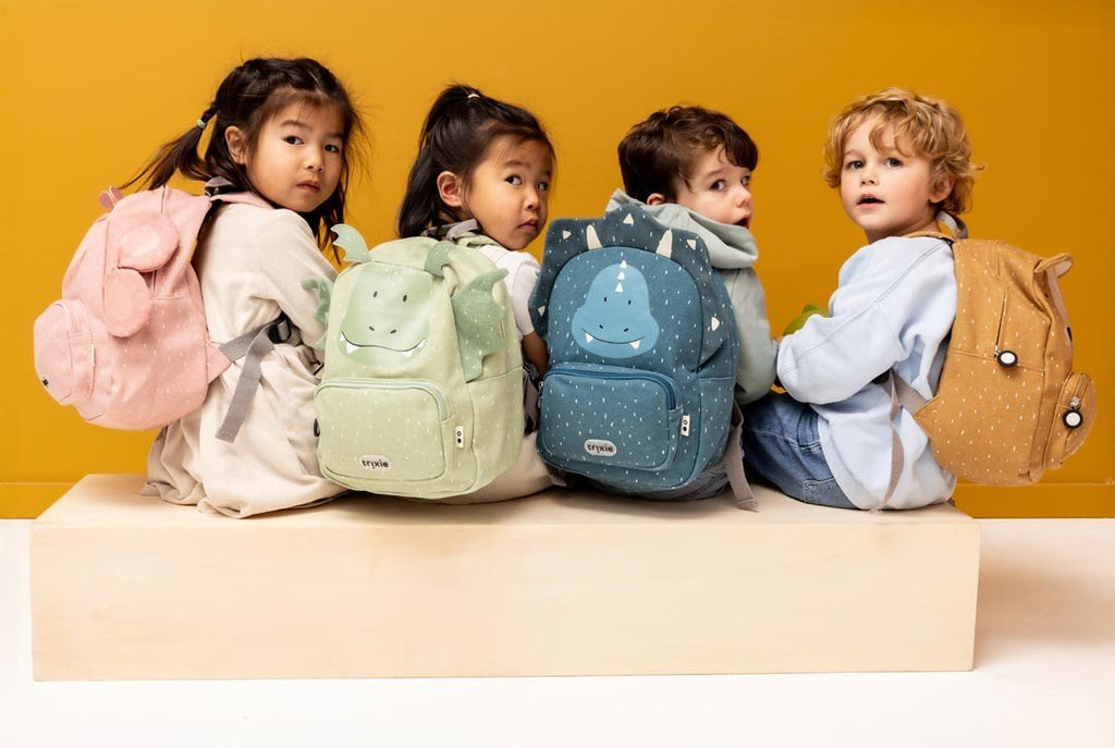 1. Four children sitting with Trixie Baby backpacks in various colors, including Mr. Triceratops and Mr. Bear, against a yellow background
