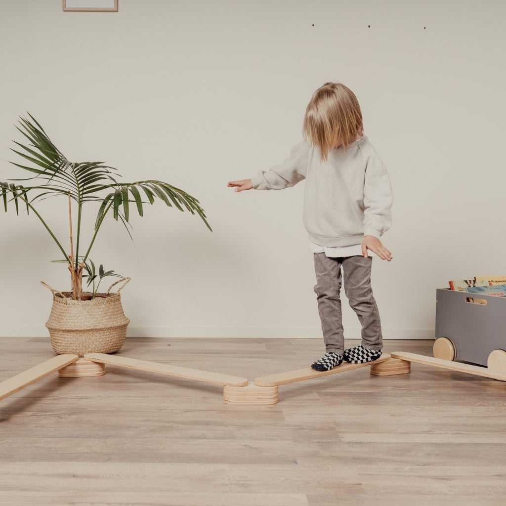 1. Child balancing on wooden balance beams in a living room setting with a potted plant and toy storage in the background