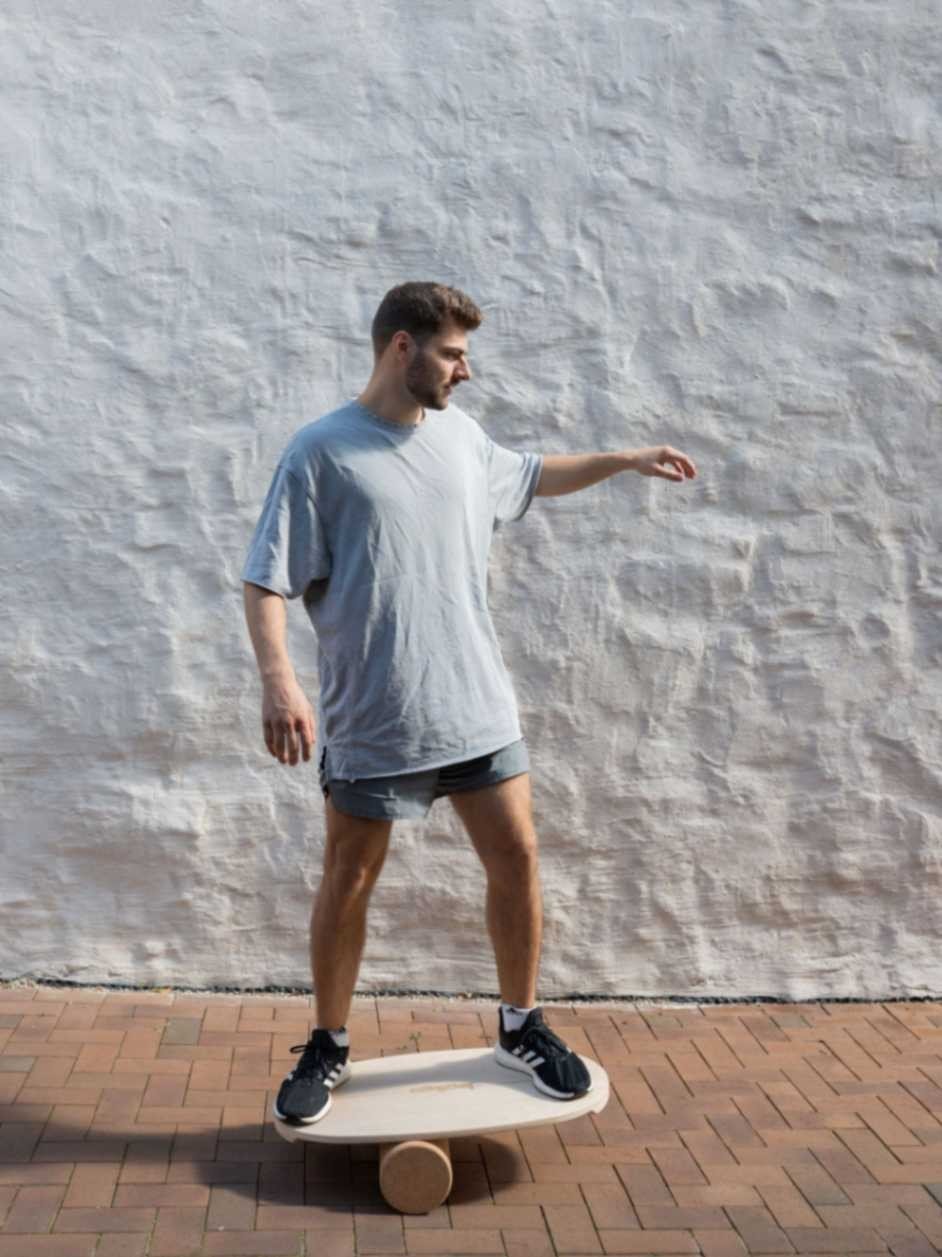 1. Man balancing on leg&go board with roller against white textured wall, wearing grey shirt and shorts