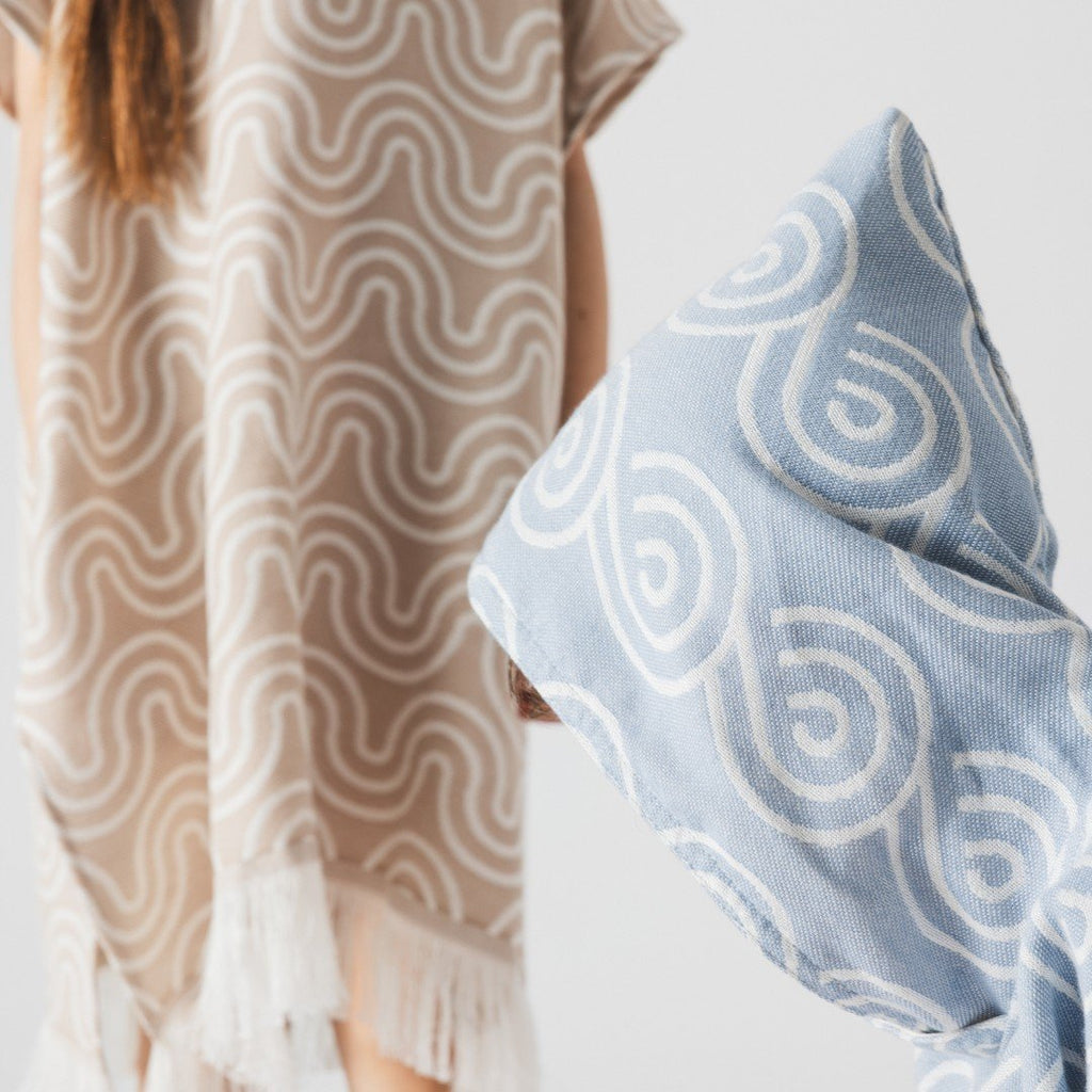 4. Close-up of sand-colored bamboo poncho with wave pattern and fringe detail next to blue poncho