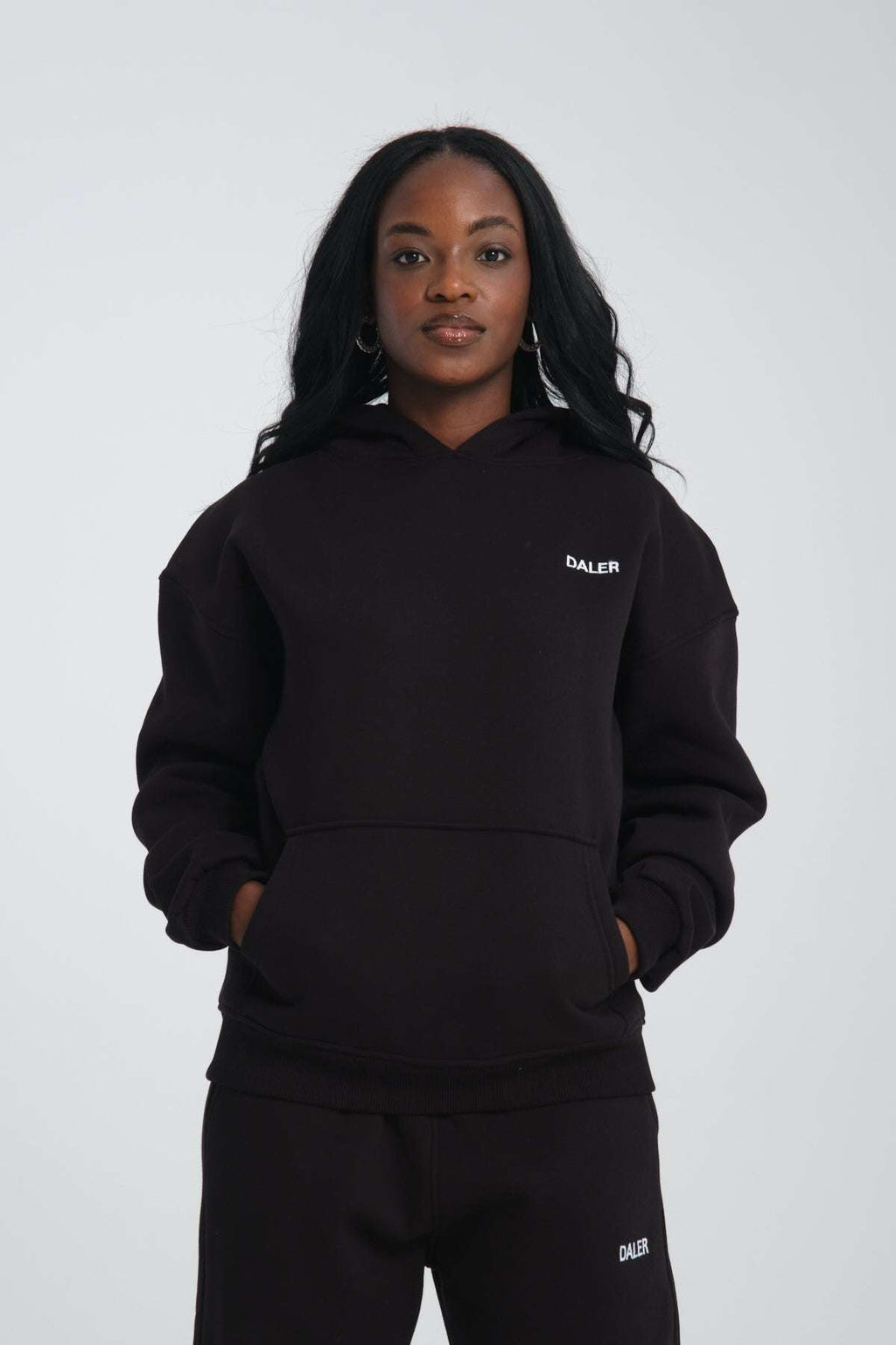 1. Woman wearing black oversized hoodie with DALER embroidery on chest, hands in pockets, studio setting