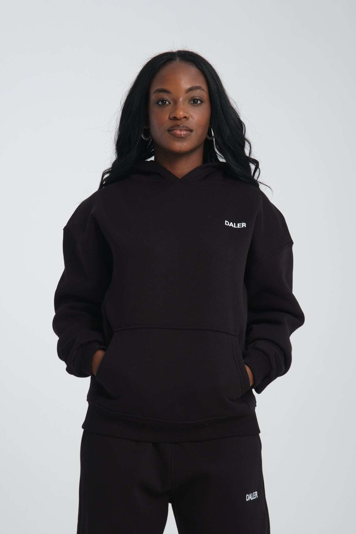 1. Woman wearing black oversized hoodie with DALER embroidery on chest, hands in pockets, studio setting