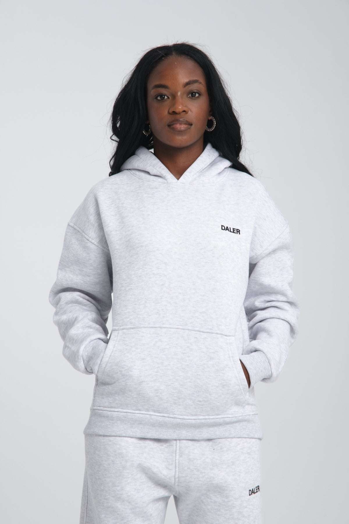 1. Woman wearing grey melange oversized hoodie with DALER embroidery, hands in pockets, studio setting