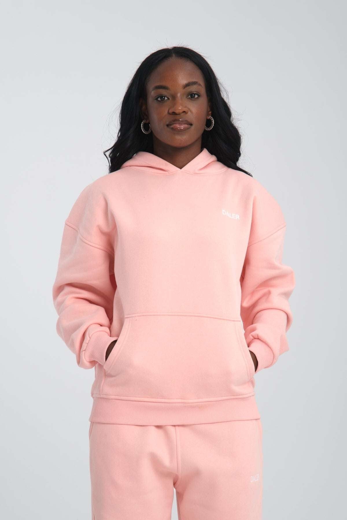 1. Woman wearing pink oversized hoodie with DALER embroidery, hands in pockets, standing against a plain background