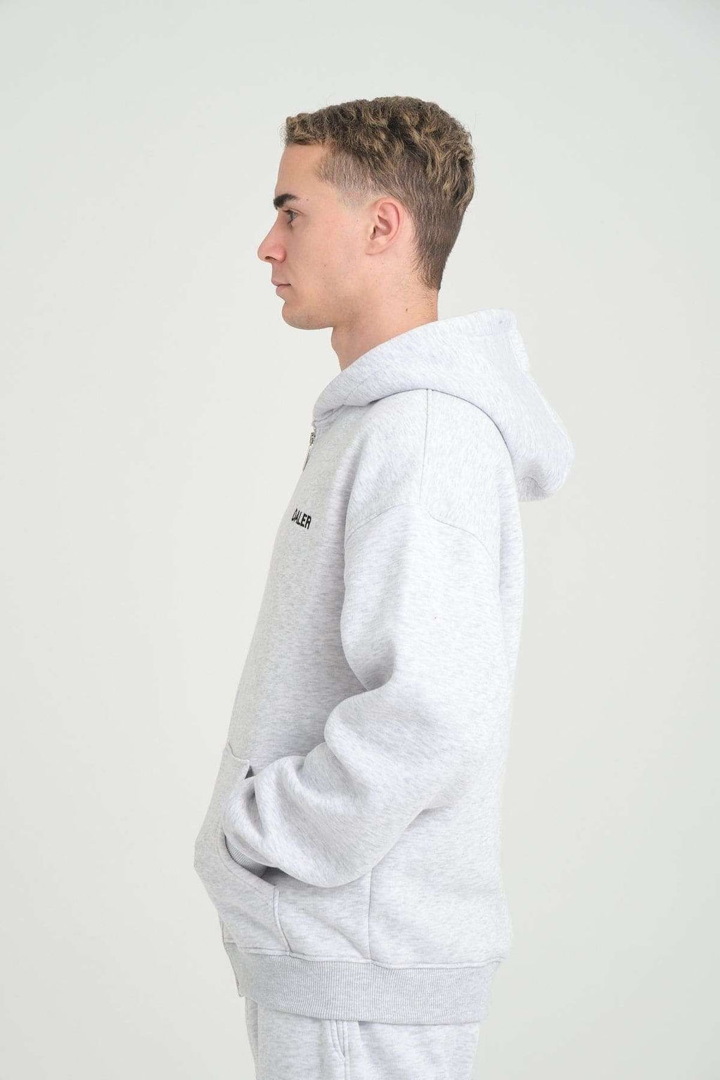 2. Side profile of man wearing grey melange DalerClo hoodie, hands in pockets, studio background