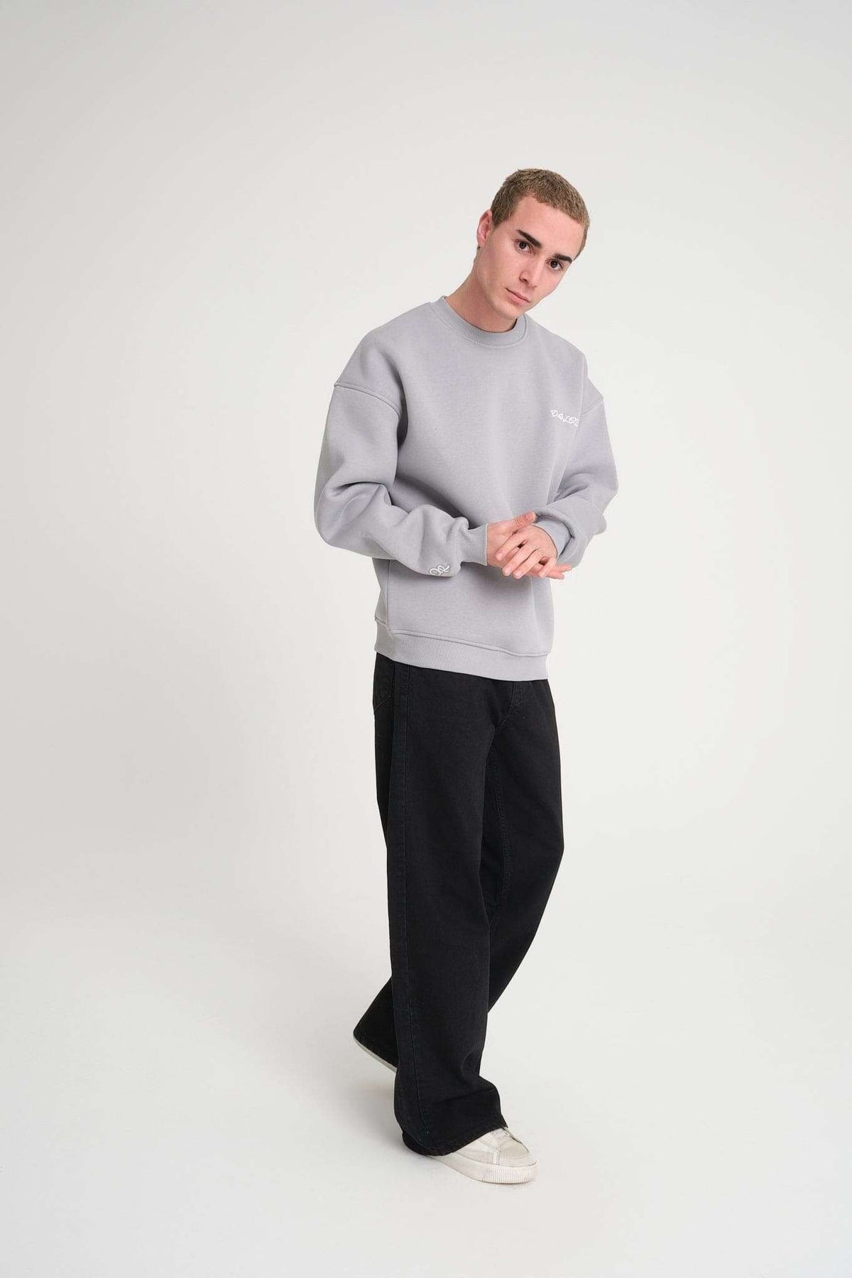 1. Man posing in grey oversized DalerClo sweatshirt with black pants and white sneakers in studio setting